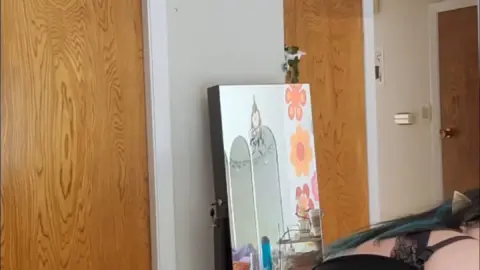 Dark-haired tattooed elf girl bends over bed with string lights