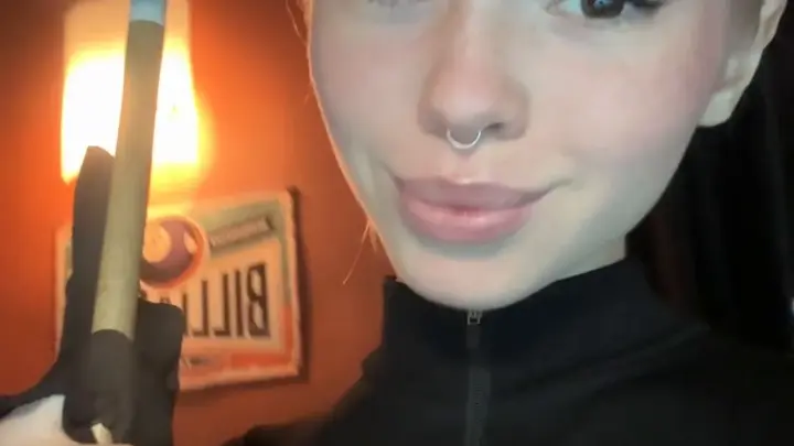 Blonde with nose ring plays pool, smiles at camera in dim bar