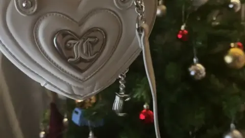 Girl holding white heart-shaped purse by Christmas tree in daylight